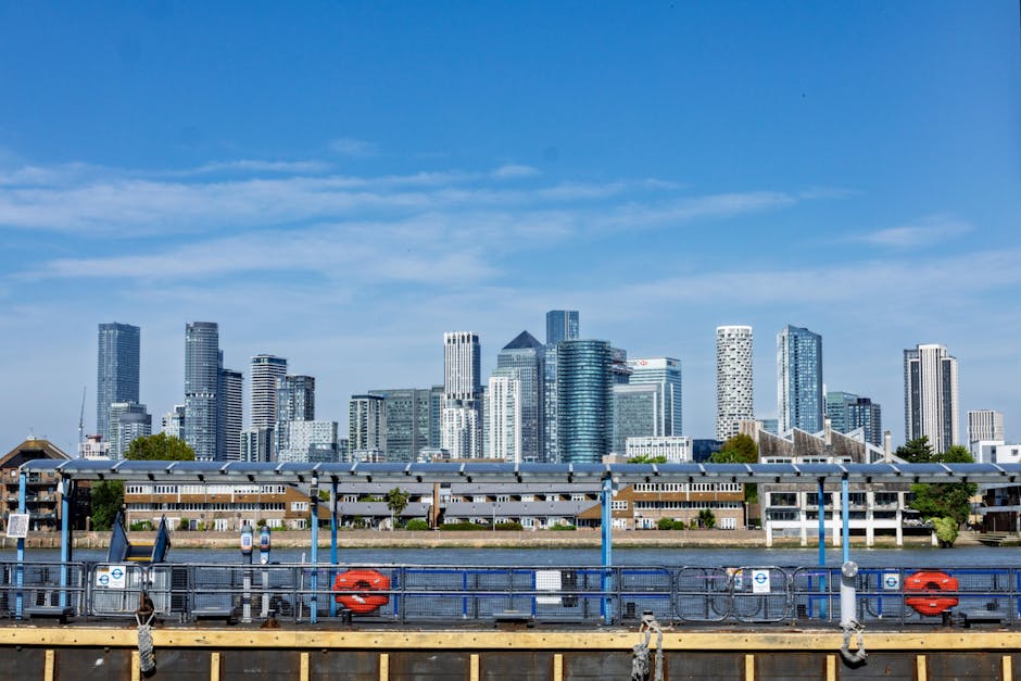A wide-angle view of a city skyline along a river at dusk, featuring modern high-rise office buildings with glass facades and illuminated windows, some with distinctive architectural designs, standing
