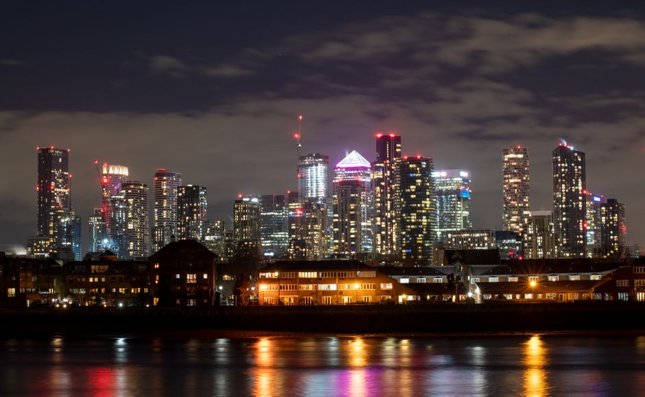A nighttime view of the Canary Wharf skyline showing tall commercial and residential skyscrapers illuminated with bright lights, including red, white, and purple accents, standing behind smaller build