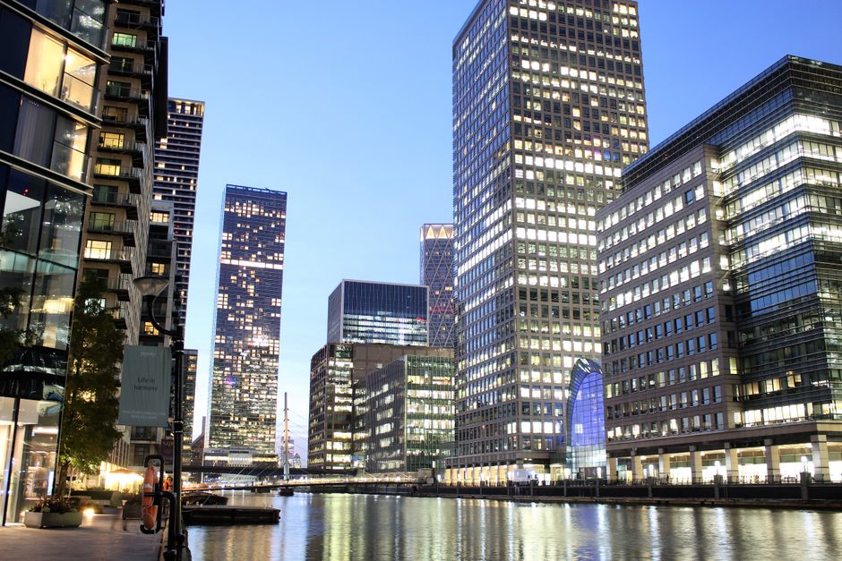 A modern multi-storey office building with a glass exterior featuring curved corners and horizontal window lines, part of a cluster of tall commercial skyscrapers in an urban setting near Canary Wharf