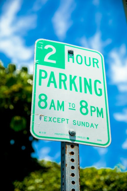A close-up of a green and white parking sign indicating a two-hour parking limit from 8 am to 8 pm, except on Sundays, mounted on a metal pole with visible rust spots. In the background, partially obs