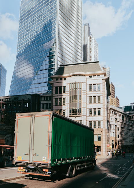 A large green delivery truck with a closed cargo area is parked on a city street in front of modern glass skyscrapers and older brick buildings, during daylight. The truck is situated close to the pav
